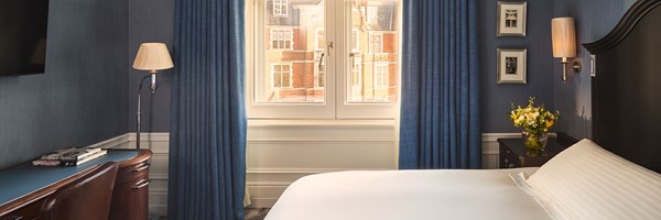 Bedroom with a white bed, blue curtains and a desk with books beside a window overlooking red-brick buildings.