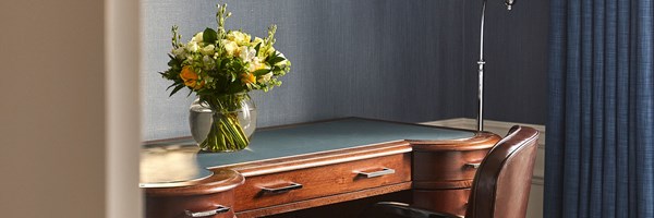 A wooden desk with a leather chair, desk lamp and a vase of flowers against blue textured walls.