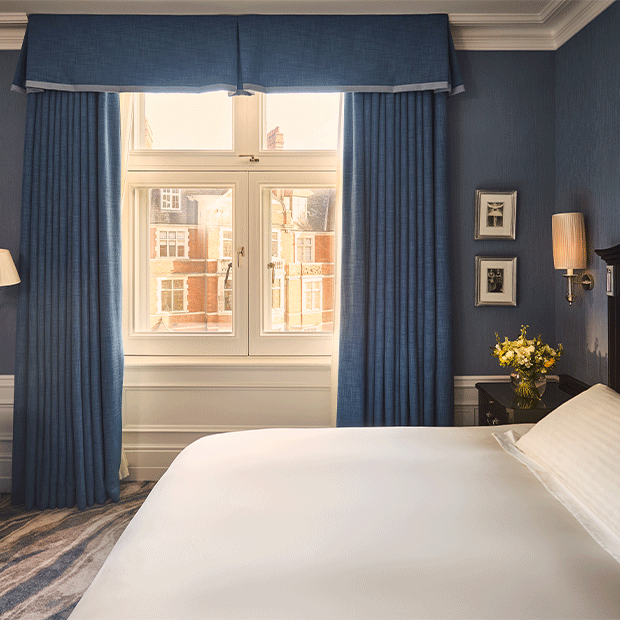A bright bedroom with blue curtains, a white bed and views of red-brick buildings through a large window.