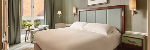 Cosy bedroom with a king bed, pale green padded headboard, matching curtains, and a window-side table with flowers and an armchair.