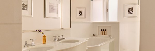 Light-filled bathroom with twin sinks, chrome fixtures, and a bathtub, decorated with framed black-and-white photographs.