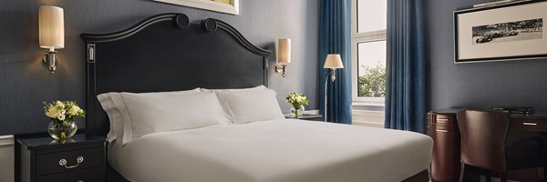 A serene hotel room with elegant dark wood furnishings, a plush bed with crisp white linens, and a window draped in rich blue curtains, accented by modern wall sconces and tasteful artwork.