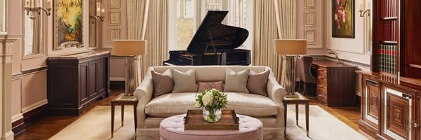 Expansive view of a classically styled drawing room with a grand piano, pink ottoman, cream sofa, symmetrical side tables with gold lamps, and walls adorned with art and mouldings.