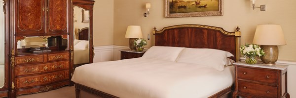 Elegant bedroom with a carved wooden headboard, crisp white bed linen, mirrored antique armoire, bedside tables with floral arrangements, and wall-mounted artwork above the bed.
