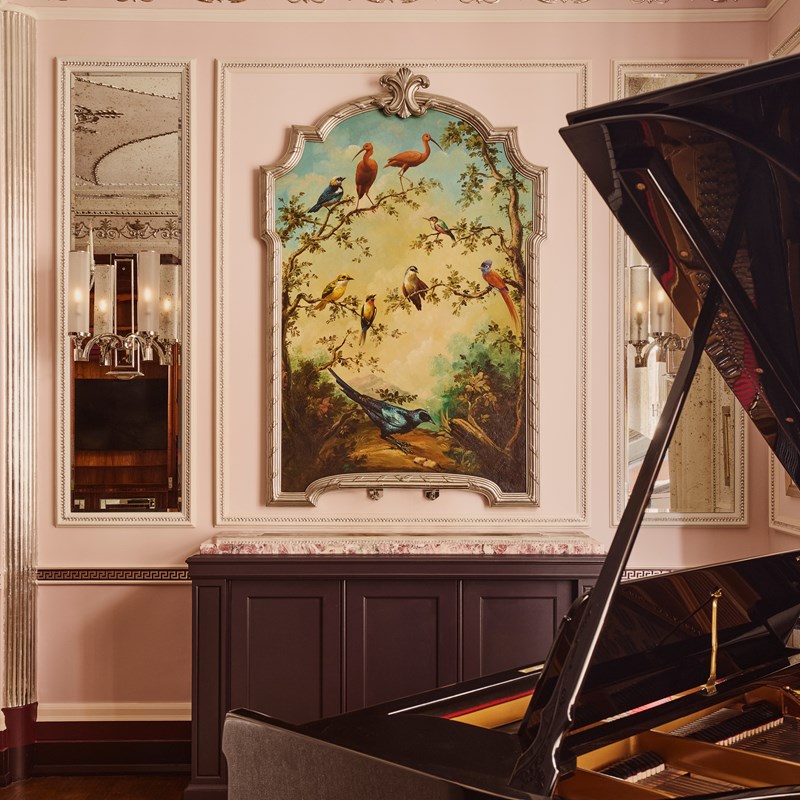 Close-up of a wall mural above a grand piano, depicting vibrant birds perched on flowering branches beneath a soft sky, framed in decorative plasterwork.