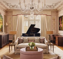 A grand salon with ornate ceiling details, a crystal chandelier, a black grand piano in front of tall curtained windows, and cream sofas with pastel cushions surrounding a pink tufted ottoman.
