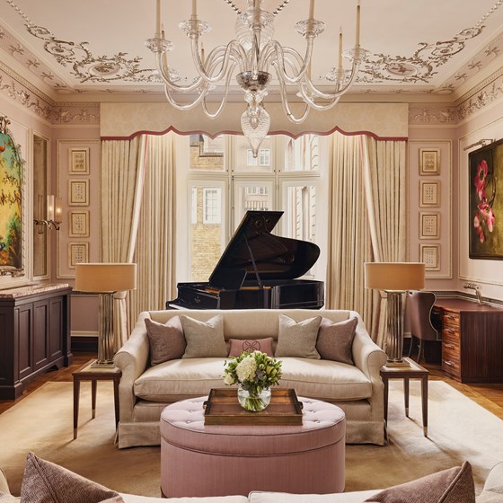 A grand salon with ornate ceiling details, a crystal chandelier, a black grand piano in front of tall curtained windows, and cream sofas with pastel cushions surrounding a pink tufted ottoman.