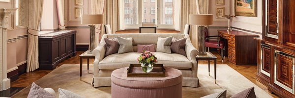 Grand salon with a cream sofa and blush ottoman, framed by tall windows, dark wood cabinetry, and rich decorative details including artwork and polished furniture.