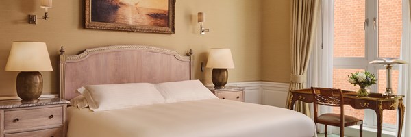 Elegant bedroom with a sculpted wood headboard, twin marble-topped nightstands with table lamps, and a classic desk positioned in front of large windows with heavy drapes.