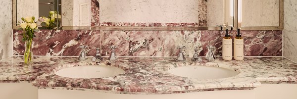 Luxurious bathroom vanity with a dual sink carved from purple-veined marble, polished chrome taps, wall mirror, and floral arrangement beside curated toiletries.