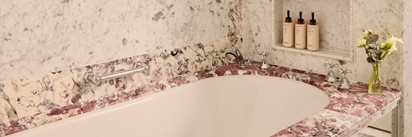 Marble-clad bathtub with intricate purple and cream veining, chrome fittings, a glass vase with white lilies, and a built-in shelf holding bath products.