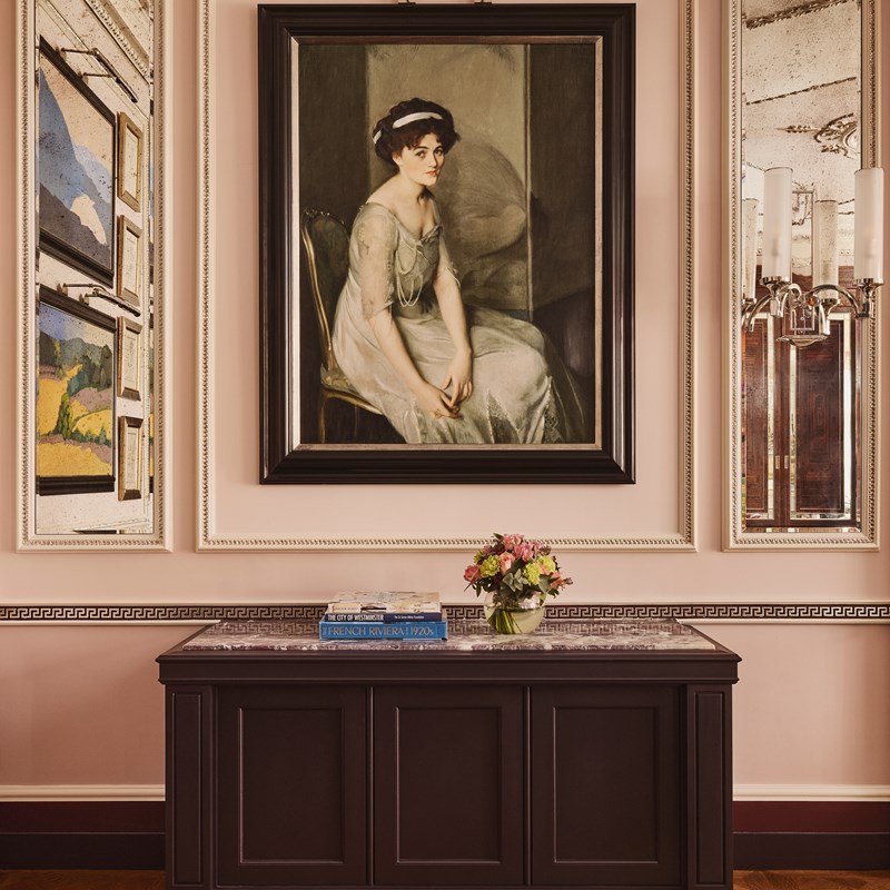 Classic portrait of a seated woman in a pale gown with a soft expression, framed in black and hung above a dark wood console with flowers and books.