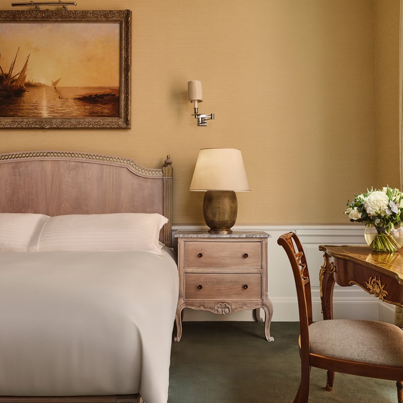 Serene bedroom corner with a pale wood headboard, marble-topped nightstand, gold-toned lamp, and a traditional writing desk adorned with fresh flowers.