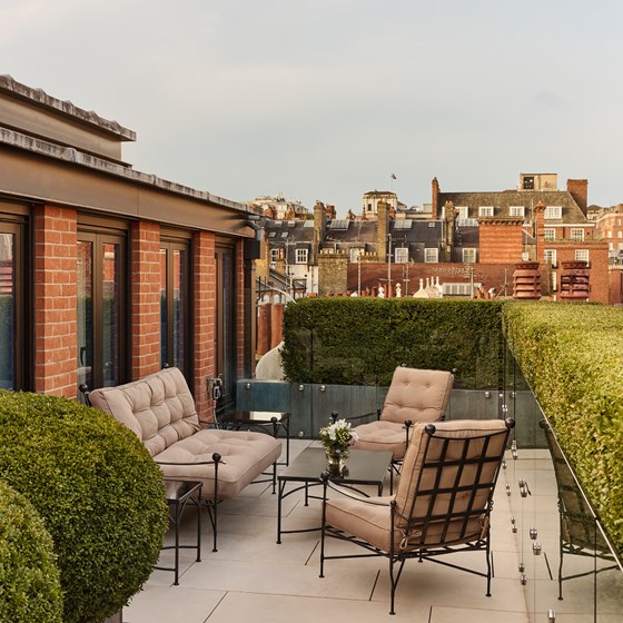 Detail of The Connaught’s Terrace Suite, showing an intimate outdoor seating area with cushioned chairs, glass balustrade, manicured hedges, and views over Mayfair rooftops.