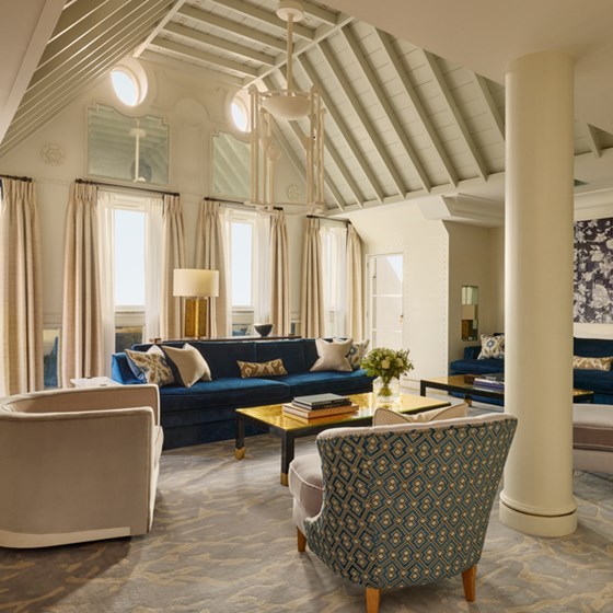 Striking vaulted-ceiling living room with rich blue velvet sofas, patterned armchairs, and golden accents, framed by tall windows and soft neutral drapery