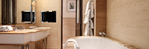 Light marble bathroom with a deep oval tub, robes on a heated rail and dual mirrors above the sink.