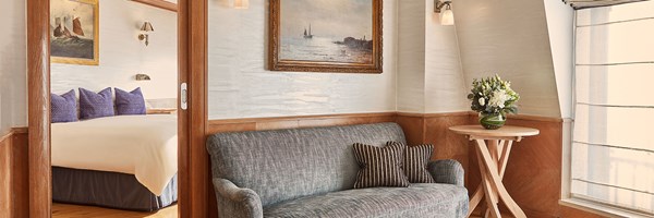 Bright sitting area with a grey sofa and artwork, opening to a bedroom with blue pillows and sailboat art.