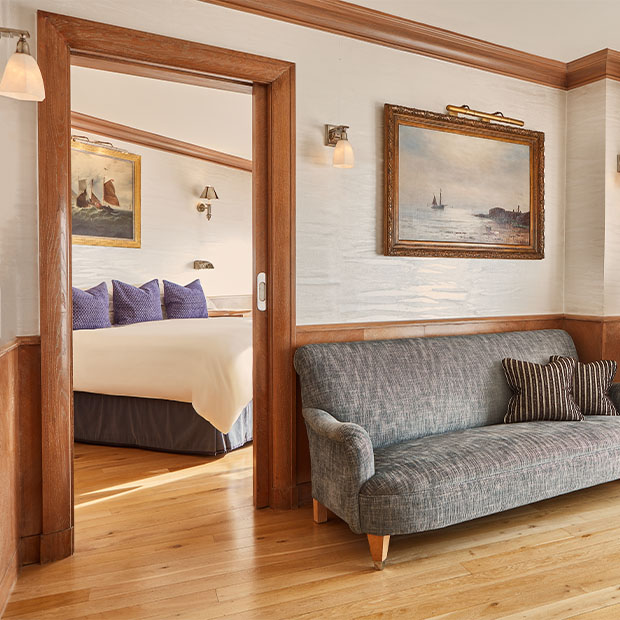 Bright sitting area with a grey sofa, leading to a bedroom with nautical art and a king bed.