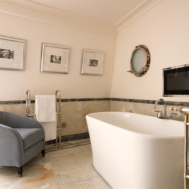Cream bathroom with a freestanding tub, blue armchair and chrome fittings beneath framed art.