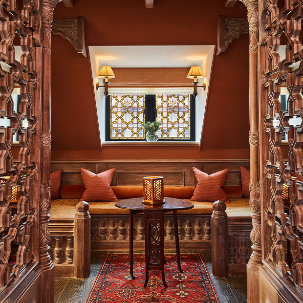 Wood-carved sitting alcove with cushions and a small lantern table beneath patterned windows.