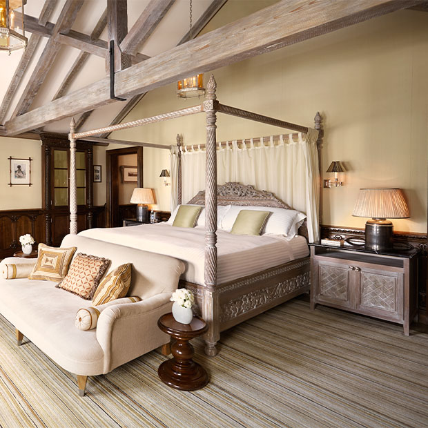 Large four-poster bed with carved posts, soft neutral linens and exposed timber beams above.