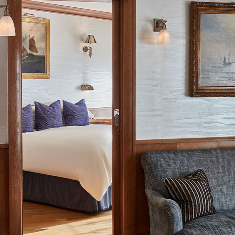 View through wooden sliding doors into a bedroom with a neatly made double bed, deep blue cushions, and maritime-themed paintings on the walls. A grey armchair and wall sconce appear in the foreground.