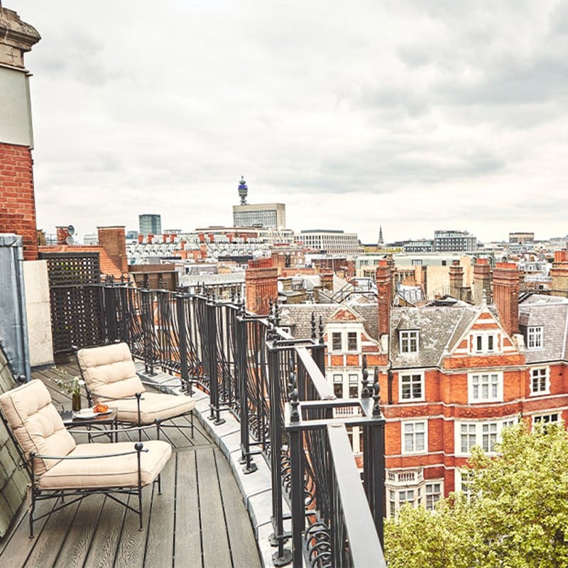 Private balcony featuring two cushioned lounge chairs, black wrought-iron railings, and panoramic views over red-brick rooftops and London landmarks under a cloudy sky.