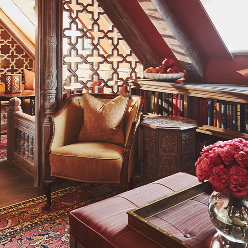 Cozy reading nook with an upholstered armchair, wooden lattice screen, carved furnishings, a bowl of fruit, shelves lined with books, and warm red and gold tones throughout.