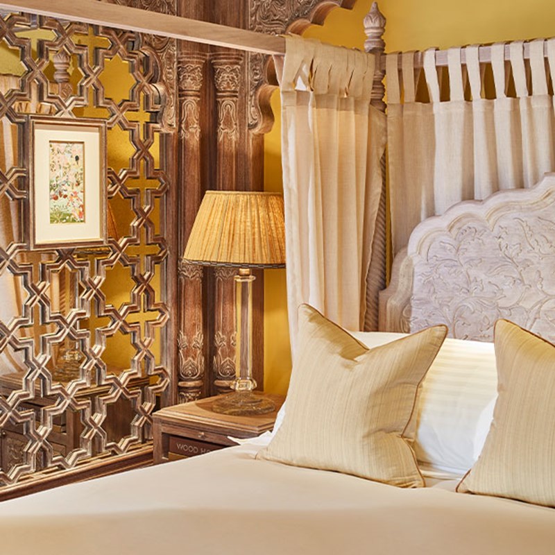 Close-up of a carved wooden four-poster bed with pale yellow cushions, an embroidered headboard, and a bedside table with an antique-style lamp framed by a decorative wood panel.