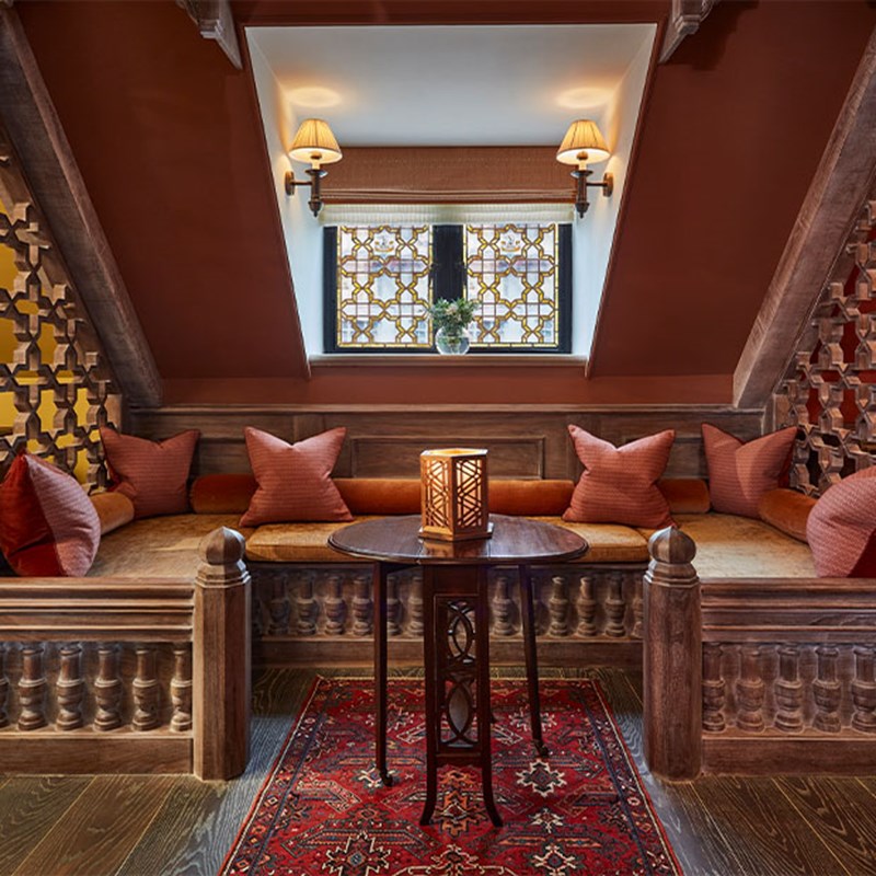 Built-in wooden bench with coral-toned cushions nestled under a sloped ceiling, intricate lattice screens, wall sconces, and a candle lantern resting on a dark wooden table atop a Persian rug.