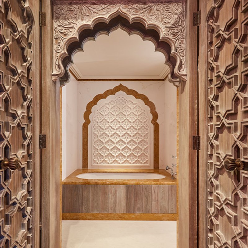 Luxurious marble-accented bathroom with an inset soaking tub framed by an elaborately carved wooden arch and patterned wall inlay, viewed through double lattice doors.