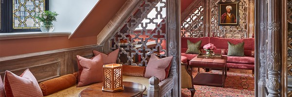 Lounge area featuring deep red velvet sofas, carved wooden lattice panels, and a portrait of a turbaned figure. Decorative pillows and a tray of flowers rest on the central ottoman, with ambient lighting and stained-glass windows.