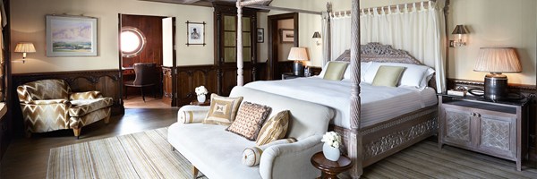 Spacious suite with a carved wooden four-poster bed, cream upholstered bench, and seating area with patterned armchair. Rich wooden panelling and a circular porthole-style window add nautical character.