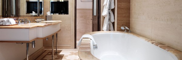 Luxurious bathroom with a curved bathtub clad in light wood, twin sinks with marble countertops, wall-mounted mirrors, and a built-in television. Neutral tones and plush bathrobes complete the setting.