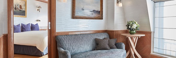 View from the sitting area with a grey upholstered sofa and nautical artwork toward a separate bedroom with a double bed and navy cushions. Soft lighting and a vase of fresh flowers enhance the tranquil ambience.