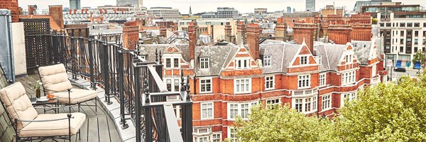 Private balcony overlooking a red-brick London streetscape, furnished with cushioned lounge chairs and a small table set with drinks and flowers. Black wrought-iron railings run along the curved terrace.