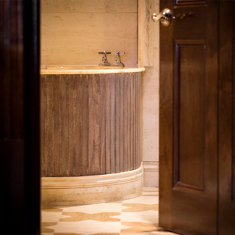 Close-up of a freestanding bathtub with ribbed wooden cladding and polished stone rim, set in a marble bathroom and partially visible through an open wooden door.