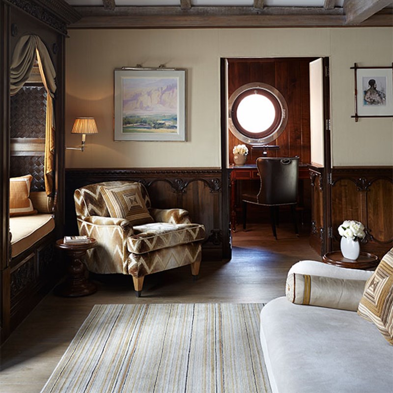 Living space with a patterned armchair, window seat, and writing nook framed by dark wood and a round porthole-style window. Soft lighting and artwork contribute to the refined, heritage feel.