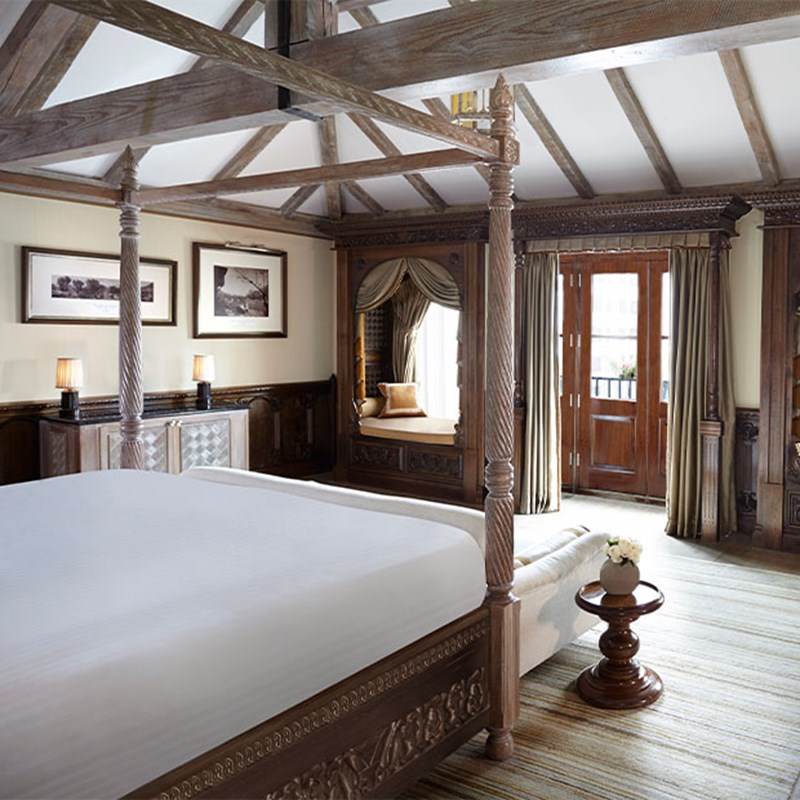 Bright bedroom with a vaulted ceiling, carved wooden four-poster bed, and French doors opening onto a private balcony. A canopied window seat and exposed beams lend rustic charm.