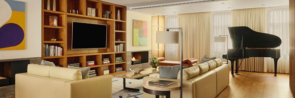 Contemporary living room with cream sofas, a grand piano, open shelving, and abstract artwork, bathed in natural light from full-height curtains.