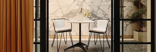 Elegant outdoor terrace with a marble backdrop, bistro-style seating, and champagne set for two, framed by floor-to-ceiling doors.