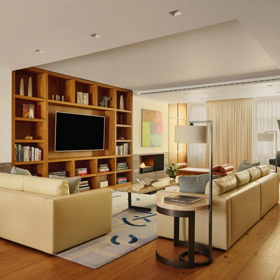 Interior view of The Mews suite, showcasing a contemporary open-plan living room with cream leather sofas, a bespoke wooden bookshelf, abstract artwork, and soft ambient lighting.