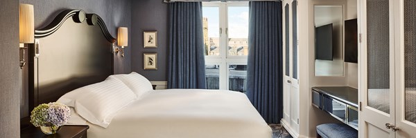 A softly lit bedroom with a dark wood headboard, crisp white bed linens and blue drapes framing a window overlooking Mayfair rooftops, with framed artwork and wall sconces adding a classic touch.