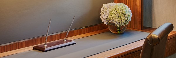 A writing desk in rich wood tones with a leather chair, two elegant pens and a vase of pale hydrangeas, creating a serene workspace.