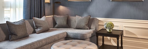A cosy sitting area featuring a corner grey sofa with textured cushions, a round tufted ottoman, and soft lighting against blue walls with framed artwork.