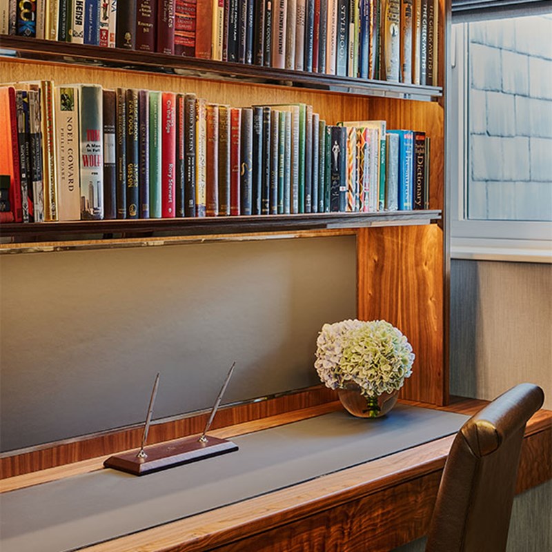 Compact study area with built-in bookshelves filled with colourful titles, a wooden writing desk, leather chair and a vase of hydrangeas.