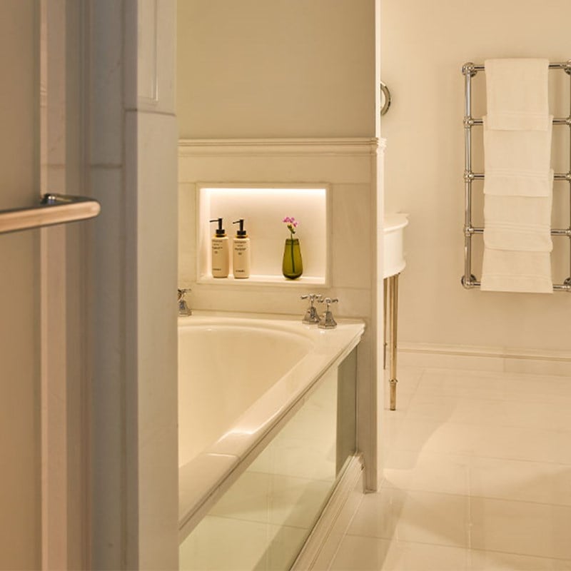 Elegant bathroom with a deep bathtub, white tiled walls, built-in shelf holding toiletries and a small green vase, and a heated towel rail.