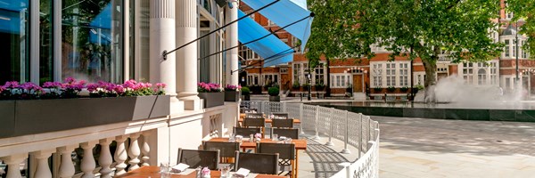 Jean-Georges at The Connaught Terrace with the sun shining down on the tables and chairs. Blue awnings cover the terrace, with flowers on the window sills and a tree to the right hand side on the pavement